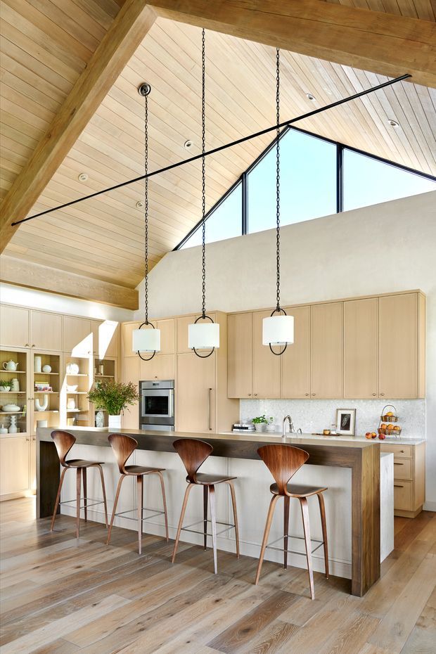 Bright open-plan kitchen with a large island and four timber bar stools, pale timber cabinetry, a light stone-look splashback, integrated stainless-steel appliances, and three suspended pendant lights. The room has a dramatic pitched timber-lined ceiling with exposed heavy timber beams, high clerestory glazing, and warm timber flooring.