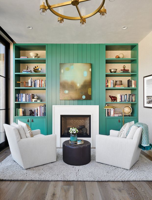 A refined living room centered on a fireplace with symmetrical built-in teal-green shelving and cabinetry. The shelves display books and decorative objects, while two light upholstered armchairs face a small dark round ottoman table. A colorful abstract artwork hangs above the white fireplace surround. The room has a textured light rug over timber flooring, large black-framed glazing at left, and a brass circular pendant light. The overall look is polished, colourful, and contemporary with classic detailing.