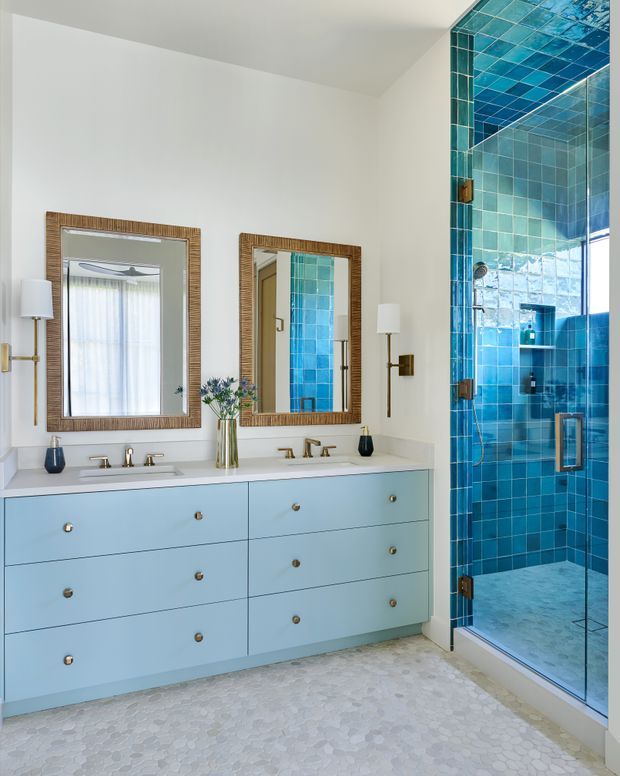 Bright, refined bathroom with a wide pale blue double vanity, white benchtop, twin framed mirrors, wall sconces, and a frameless glass shower enclosure lined in vivid turquoise-blue tiles. The floor is finished in small light stone-look mosaic tiles, creating a fresh coastal feel.