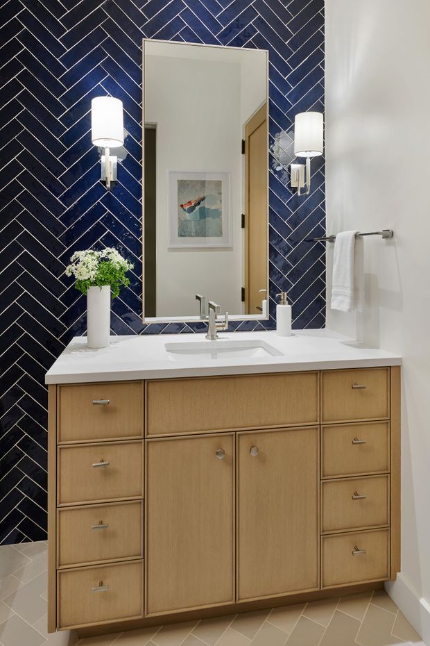A compact, refined bathroom vanity area with a large rectangular mirror, white integrated basin and benchtop, and extensive light timber cabinetry. The main feature is a deep navy-blue ceramic tile wall laid in a herringbone pattern with contrasting light grout. Two modern wall sconces flank the mirror, while a white towel hangs on a chrome rail and a small plant sits on the vanity. The floor is finished in light beige geometric tiles.