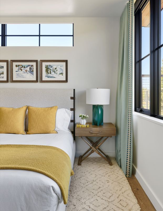 A bright, compact bedroom featuring a neatly made bed with white linens, mustard-yellow pillows and throw, a textured upholstered headboard, and a wood bedside table with a teal lamp. Framed artwork hangs above the bed, while large black-framed windows with pale green curtains provide abundant natural light. Light wood flooring and a textured cream rug create a warm, inviting feel.