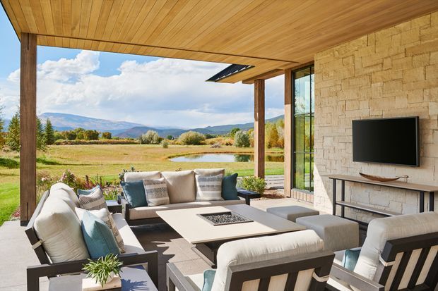 A spacious covered outdoor living area with a warm timber-lined ceiling, timber posts, and a pale natural stone feature wall. The space is furnished with a large cream sectional sofa, cushioned lounge chairs, a low rectangular coffee table with a fire feature, and a wall-mounted television. Full-height glazing opens the room to expansive rural views and distant mountains, creating a relaxed indoor-outdoor entertaining setting.