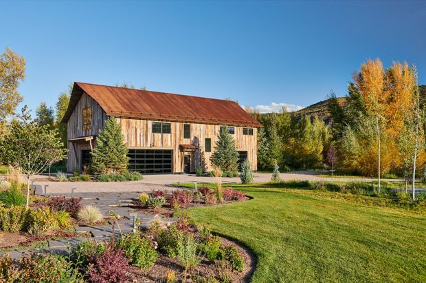 Exterior view of a rustic timber-clad barn-style building with a weathered red metal gable roof, large garage door, and landscaped grounds. Autumn trees and a mountainous backdrop surround the property, with a paved garden path and planted beds in the foreground.