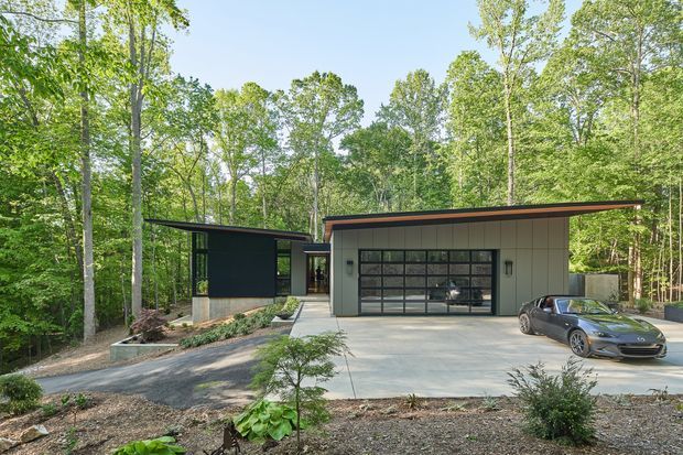 A contemporary single-storey residence or ancillary building set within a wooded landscape. The composition shows a broad concrete driveway and garage frontage with a dark-framed glazed garage door, pale vertical wall cladding, and a low asymmetrical roof with a warm timber-toned edge. A dark pavilion-like wing extends to the left, connected by a glazed link. Mature deciduous trees surround the property, and a parked car is visible in the driveway.