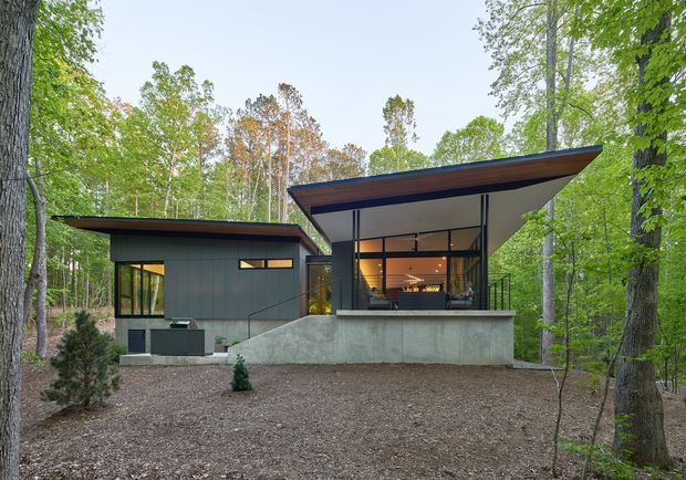 A contemporary single-storey house set among mature woodland. The composition features two connected flat or shallow mono-pitched roof volumes, dark vertical cladding, extensive floor-to-ceiling glazing, and a raised concrete terrace or plinth. Warm interior lighting is visible through the glazed living area, while the surrounding natural setting dominates the exterior view.