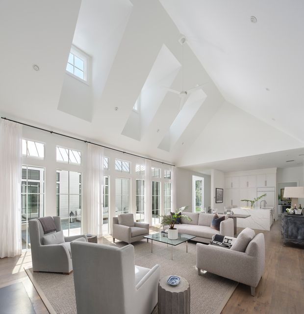 Bright, spacious open-plan living room with a dramatic vaulted ceiling, multiple skylights, clerestory-style windows and extensive floor-to-ceiling glazing. The room features neutral upholstered seating arranged around a glass coffee table, a large textured rug, white sheer curtains, white built-in kitchen cabinetry, and warm timber flooring. The overall aesthetic is refined, airy and contemporary with traditional window detailing.