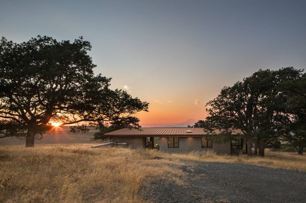 A low, single-storey modern rural residence set among mature spreading trees and dry golden grass. The compact building has a long, low profile, dark rendered walls, extensive glazed openings, and a shallow corrugated metal roof. A gravel driveway approaches the house, with warm sunset light and distant hills visible beyond.