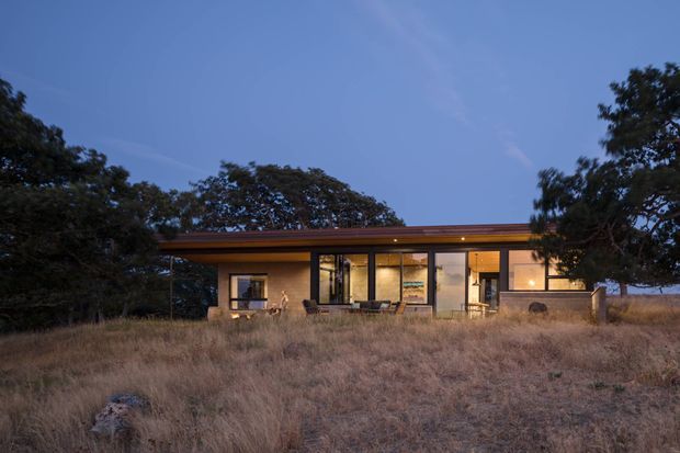 A low, single-storey modern dwelling set in a dry grassy landscape at dusk. The house has a long, shallow roof with broad eaves, extensive floor-to-ceiling glazing, dark slender framing, warm interior lighting, and a sheltered front terrace. Mature trees surround the structure, creating a calm, secluded architectural exterior view.