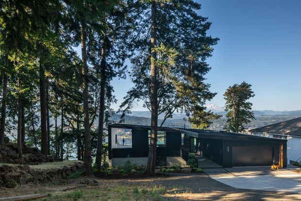 Exterior view of a low-profile contemporary dark-clad house set among tall forest trees on a sloping site. The building has a broad shallow-pitched or nearly flat roofline, large glazed openings, and an integrated double garage. A mountain and valley landscape is visible beyond the property.