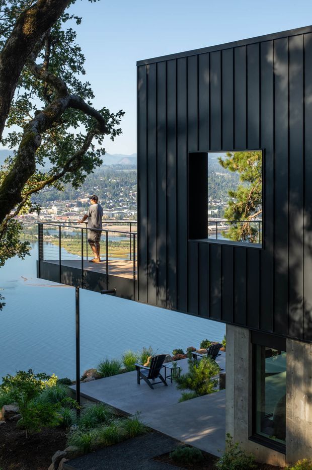 Exterior architectural photograph of a contemporary hillside home overlooking a broad waterway and urban landscape. The building features a dark, vertically ribbed metal-clad upper volume with a deep rectangular window opening, a projecting balcony with slender black railings, exposed concrete supports, and extensive glazing below. Terraced outdoor areas with concrete paving, planted beds, and outdoor lounge chairs step down the sloping site beneath mature trees.