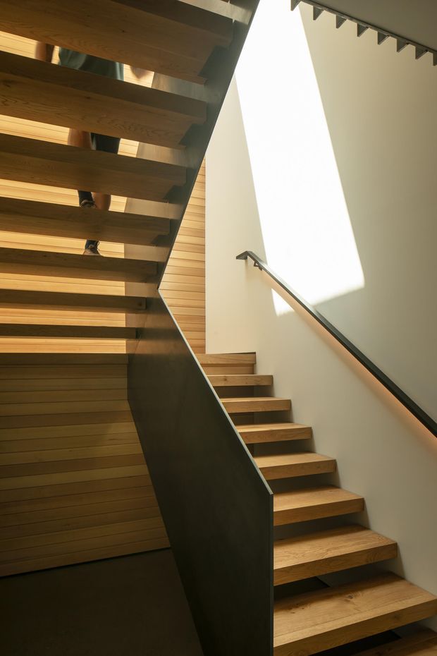 Interior architectural photograph of a contemporary stairway with open-riser timber treads, a dark metal central stringer and balustrade, warm timber wall lining, and a pale plaster wall illuminated by strong natural light. The stair rises through a compact multi-level circulation space, with a second flight visible overhead.