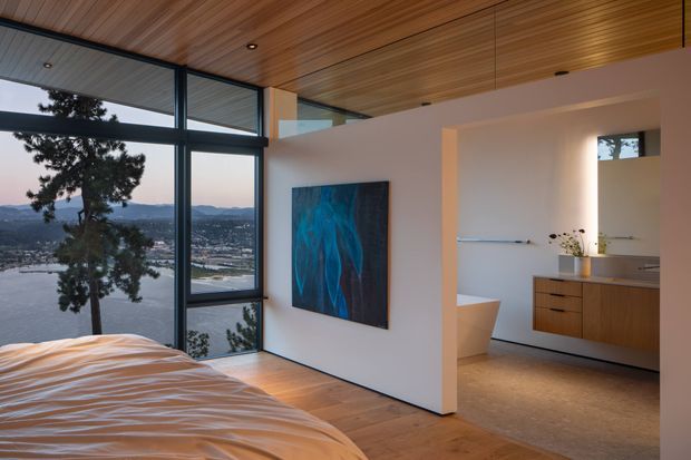 A refined bedroom with a partially visible bed, warm timber-lined ceiling, and timber flooring. Floor-to-ceiling dark-framed glazing overlooks a lake and surrounding hills. An open ensuite bathroom is visible beyond a partition, featuring a freestanding bathtub, floating timber vanity, large mirror, and pale tiled floor. A blue abstract artwork adds contrast to the otherwise warm, neutral interior.