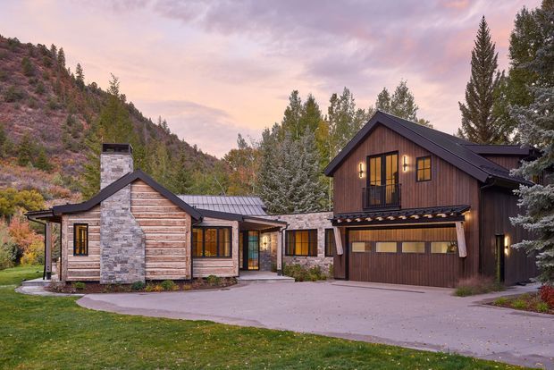 Exterior view of a mountain retreat composed of several connected, pitched-roof volumes. The home features horizontal timber siding, dark metal roofing, natural stone chimney and entry walls, warm-lit windows, and an integrated double garage. It is set against a wooded, steeply sloped mountain landscape at dusk.
