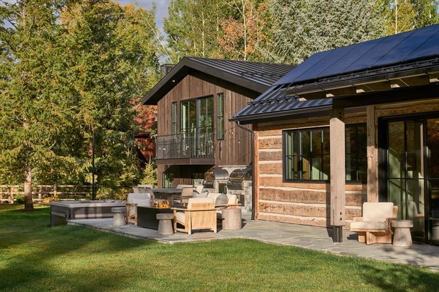 Exterior view of a contemporary timber-clad home set in a wooded landscape. The building features dark pitched metal roofs with solar panels, extensive black-framed glazing, a raised upper-level balcony, and a covered outdoor living area with built-in barbecue and lounge furniture. A paved terrace and lawn surround the house.