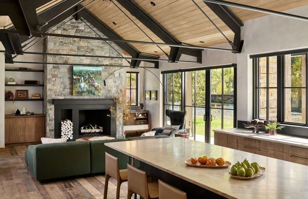 A spacious open-plan living and dining area with a large kitchen island in the foreground. The room features a vaulted timber-lined ceiling with exposed dark structural beams, extensive black-framed glazing, a natural stone fireplace wall, timber flooring, built-in shelving and cabinetry, and a contemporary seating area. The space opens visually to a landscaped outdoor area through tall glazed doors and windows.