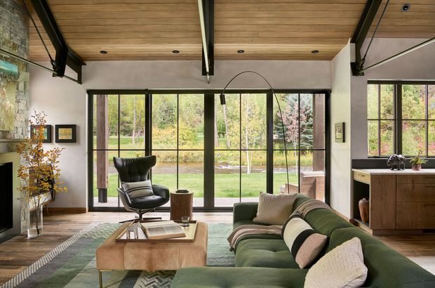 A warmly styled contemporary living room with a low green sectional sofa, leather lounge chair, and rectangular wood coffee table. Full-height black-framed glazing opens the room visually to a landscaped garden. The room has exposed timber ceiling boards with dark structural beams, pale plaster walls, timber flooring, a stone feature wall at left, and a compact timber kitchen or joinery zone at right. The overall atmosphere is warm, refined, and connected to the garden.