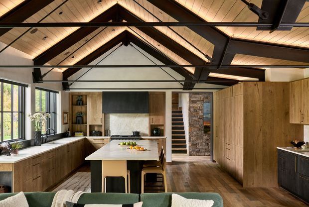 A warm, spacious contemporary kitchen with a large central island, extensive timber cabinetry, dark benchtops, and a broad bank of windows along the left wall. The room features a dramatic vaulted timber ceiling with exposed dark steel or timber beams, integrated appliances, open shelving, and a stone-clad vertical feature beside a stair opening. A partial sofa is visible in the foreground, suggesting an open-plan connection to a living area.