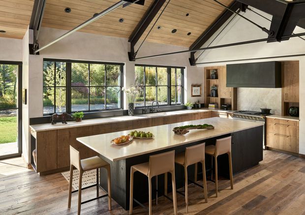 A spacious contemporary kitchen with a large central island and seating for four. The room features extensive black-framed glazing, warm timber cabinetry and flooring, a pale stone island benchtop, integrated appliances, and a dramatic exposed dark structural beam system beneath a timber-lined vaulted ceiling. Garden views are visible through the windows.