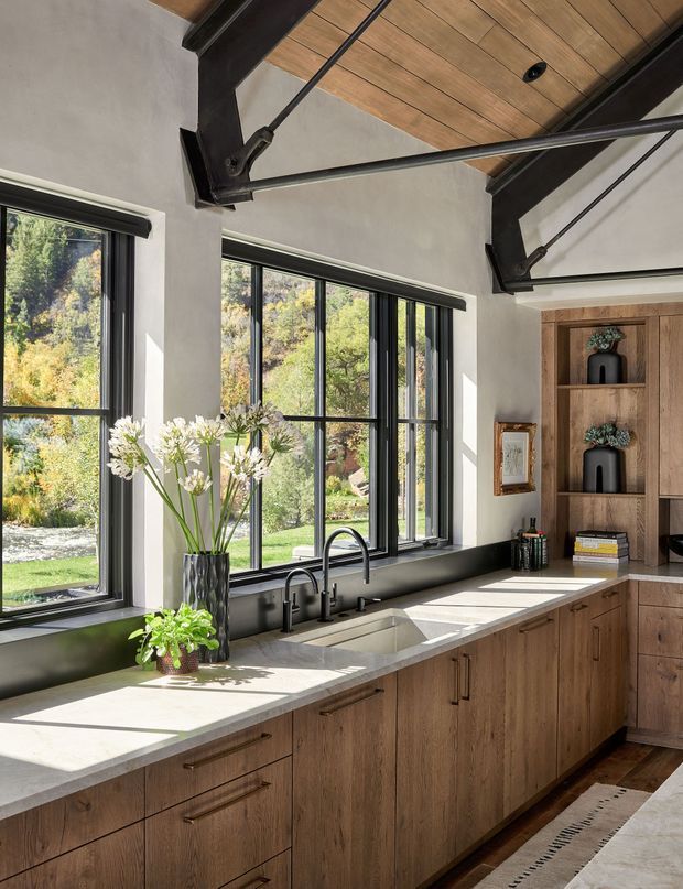 A bright, spacious kitchen with a long light stone countertop, integrated double sink, black fixtures, and extensive black-framed glazing overlooking a leafy landscape. Warm timber cabinetry and open shelving run along the wall, while exposed dark structural beams and a timber-lined ceiling add a rustic contemporary character.