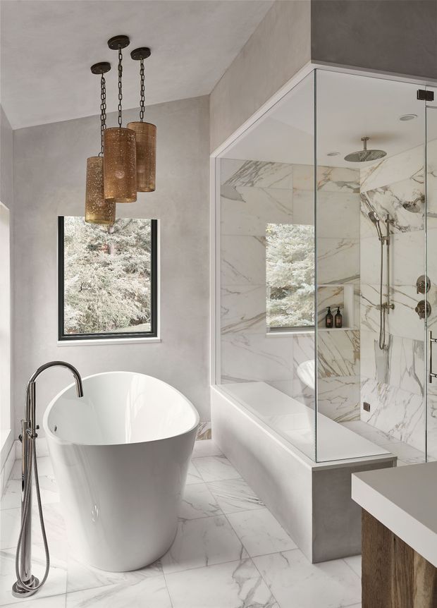 A refined contemporary bathroom featuring a freestanding white soaking bathtub, wall-mounted and handheld chrome fixtures, a large frameless glass shower enclosure, and full-height white marble-look wall and floor tiling with grey veining. The room has pale plaster walls, a dark-framed window overlooking trees, a floating vanity with a pale benchtop and timber-toned base, and three suspended bronze cylindrical pendant lights.