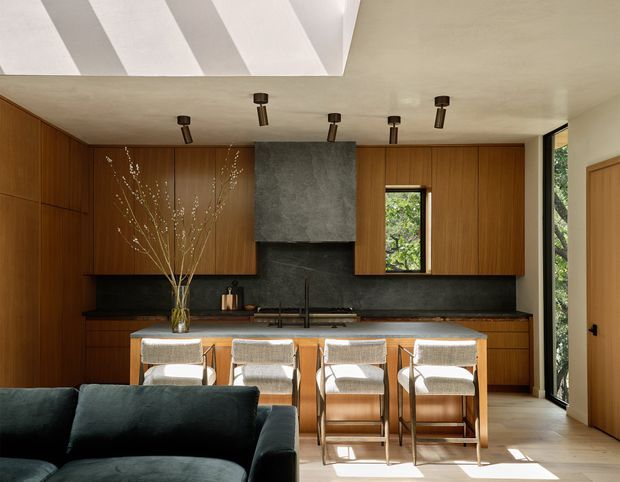 Warm contemporary open-plan kitchen and living area with extensive vertical timber cabinetry, a dark stone or concrete-look backsplash and rangehood surround, a long island with four upholstered bar stools, and a dark sofa in the foreground. Natural light enters through a narrow window and full-height glazed opening, while angled ceiling geometry and black directional spotlights add architectural interest.