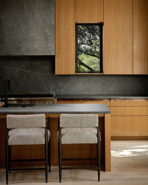 A refined modern kitchen with warm vertical timber cabinetry, a dark textured stone backsplash and benchtop, and a central island with two upholstered bar stools. A narrow window framed within the cabinetry brings in views of leafy vegetation, while pale timber flooring and natural light add warmth.