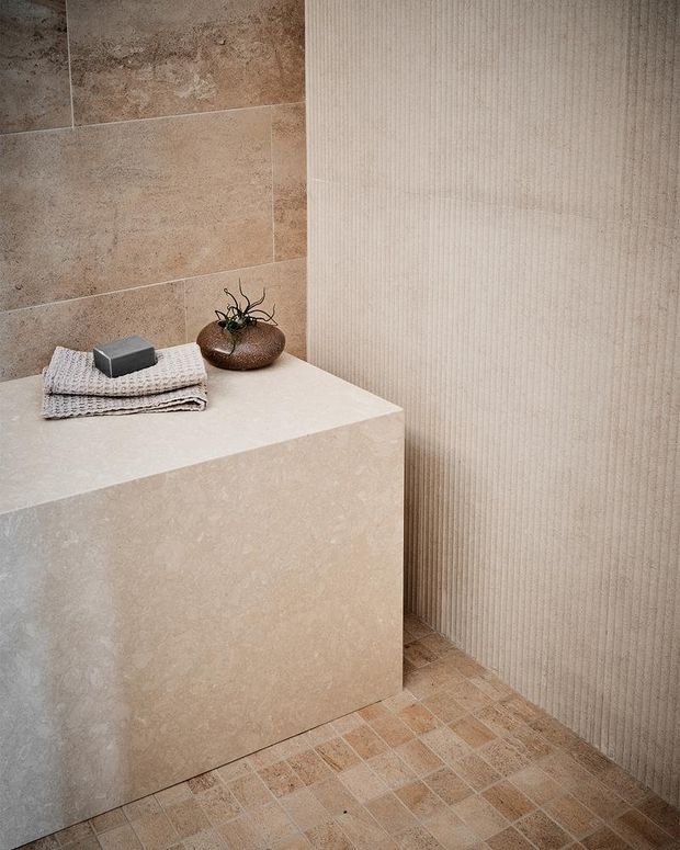 A close-up of a refined minimalist bathroom featuring a large integrated stone-clad vanity or bench, warm beige stone wall tiles, a vertically ribbed textured wall surface, and small neutral-toned accessories. The space has a calm, monochromatic palette with layered natural textures and understated detailing.