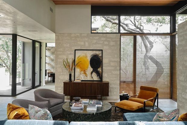 A refined contemporary living room with expansive black-framed glazing, textured pale stone or plaster walls, warm timber ceiling detailing, and horizontal timber blinds. The room features a low dark timber sideboard, abstract artwork, sculptural decor, a dark round coffee table, a grey lounge chair, a mustard velvet armchair, and a large sectional sofa in the foreground. Mature tree branches are visible outside through the glazing.
