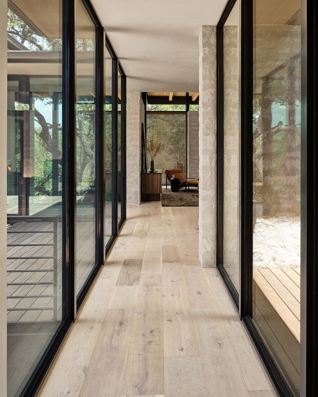 A refined contemporary interior corridor lined with extensive black-framed floor-to-ceiling glazing on both sides, connecting to a furnished living area. The hallway features pale timber flooring, textured light stone structural columns, warm timber ceiling details, and strong visual connections to landscaped outdoor decks and trees.