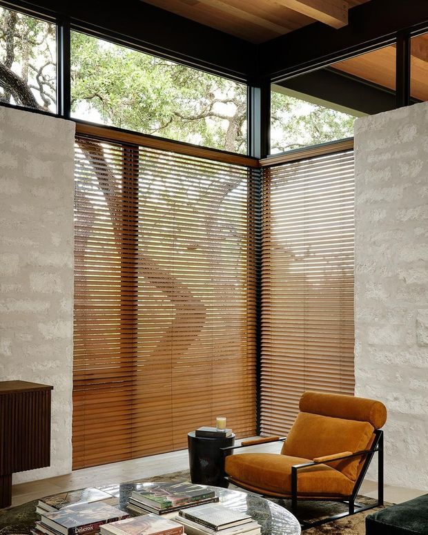 A refined interior corner with floor-to-ceiling corner glazing, warm timber venetian blinds, dark window frames, white textured masonry walls, and a timber-lined ceiling. A cognac leather lounge chair sits beside a small black side table, with books and magazines on a round coffee table in the foreground. Mature trees are visible through the glazing.
