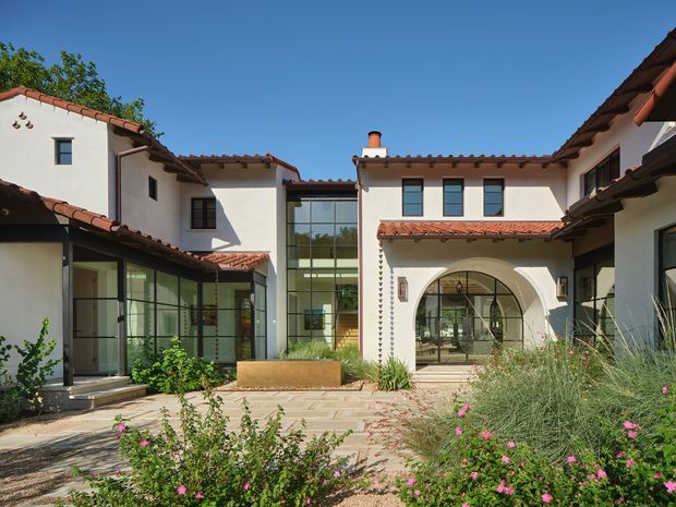 Exterior view of a large courtyard-style residence with white stucco walls, terracotta tiled roofs, dark-framed glazing, and a prominent arched entry. The paved courtyard is landscaped with flowering shrubs and ornamental grasses, with multiple connected wings and a glazed central section visible.