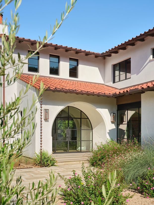 Exterior view of a two-storey courtyard residence with white stucco walls, terracotta tiled pitched roofs, dark-framed windows, and a prominent arched glazed entry. A landscaped garden and paved path occupy the foreground under clear daylight.