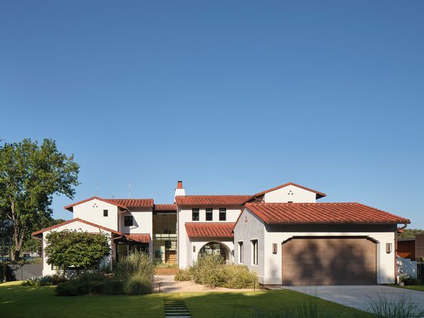 Exterior view of a large Mediterranean-inspired residential home with white stucco walls, multiple red terracotta-tiled pitched roofs, arched openings, dark-framed windows, and an attached double garage. The house is set within a landscaped suburban garden with ornamental grasses and a stepping-stone path under a clear blue sky.