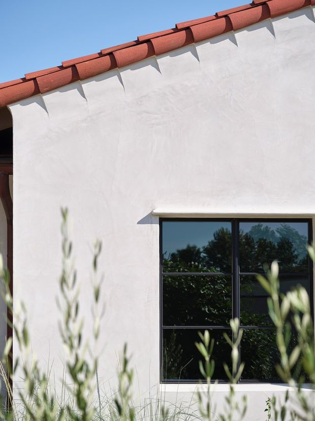 Close-up exterior view of a white rendered stucco wall with a black-framed multi-pane window and terracotta curved roof tiles. Soft-focus grasses and foliage occupy the foreground, while the window reflects trees and sky. The simple Mediterranean-influenced facade is brightly lit against a clear blue sky.