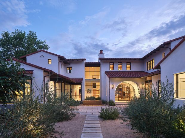 A dusk exterior view of a large courtyard-style residence arranged around a central entry garden. The building has white stucco walls, dark red clay-tile pitched roofs, timber-toned window and door frames, and a warmly illuminated glazed central stair or entry volume. A paved stepping-stone path crosses a gravelled, drought-tolerant landscaped courtyard toward an arched entrance.