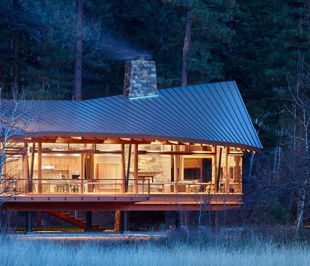 A warmly illuminated elevated cabin-style house in a dense forest at dusk. The building features a steep metal gable roof, a prominent stone chimney, extensive timber-framed glazing, exposed timber structure, and a wraparound deck with dark metal balustrades. The woodland setting and cool blue evening light contrast with the warm interior lighting.