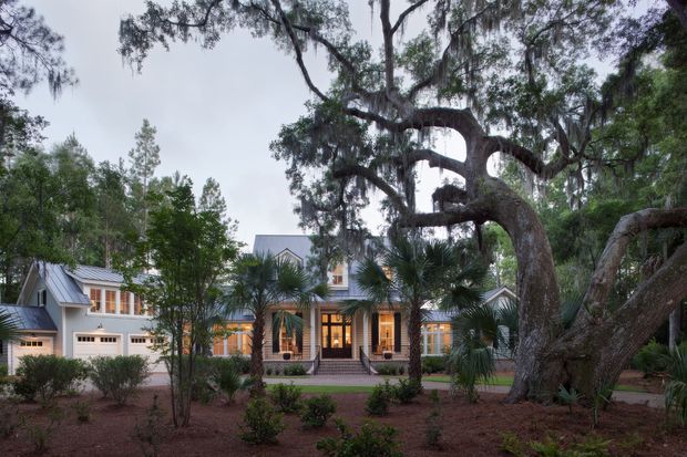 Exterior view of a large, traditional-looking house set among mature trees and landscaped planting. The facade combines light cladding, multiple gabled roof sections, a prominent central entrance, warm-lit windows, and an attached garage. A very large spreading tree dominates the foreground, with palms and low shrubs around the property.
