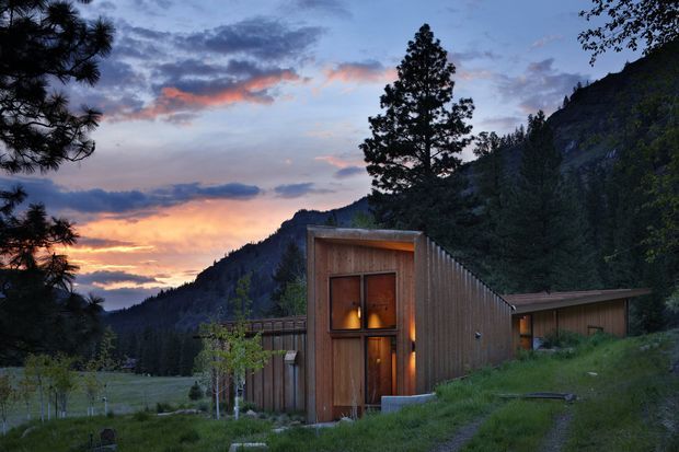 A compact modern timber-clad building set into a grassy mountain landscape at dusk. The foreground structure features a tall, recessed timber entry door framed by a weathered wood portal, with warm interior lighting visible through narrow glazing. A low, elongated wing extends to the right, while dense conifer forest, steep mountain slopes, and a dramatic sunset form the backdrop.