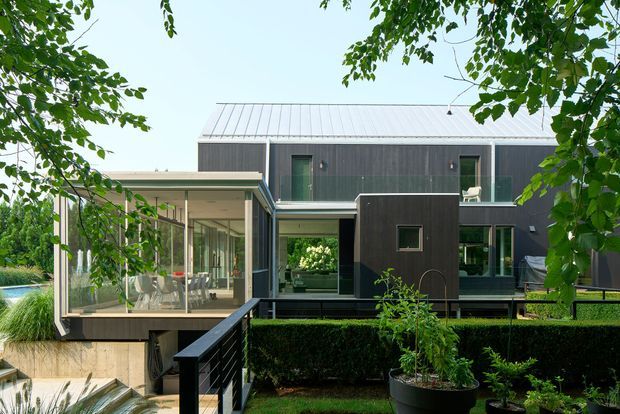 Exterior view of a contemporary two-storey residence framed by mature trees and landscaped hedges. The home features dark vertical cladding, a light grey metal pitched roof, extensive glazing, and a glazed projecting sunroom or living extension above a partially visible lower level. A stepped walkway, black balustrade, garden planting, and a glimpse of water appear in the foreground.