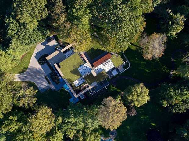 Aerial top-down view of a large contemporary residence set within dense, mature woodland. The building has a complex low-profile composition with multiple flat and planted roof sections, contrasting roof surfaces, courtyards or terraces, and a broad curved driveway approaching from the left.