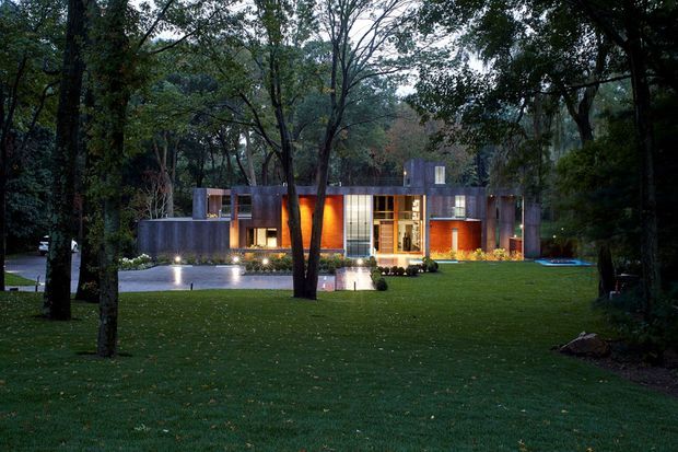 A low, modernist flat-roofed house set within a mature wooded landscape. The façade combines dark grey architectural volumes, extensive floor-to-ceiling glazing, and warm timber-toned illuminated feature walls. A broad manicured lawn, reflecting pool or water feature, and subtle exterior lighting create a calm evening setting.