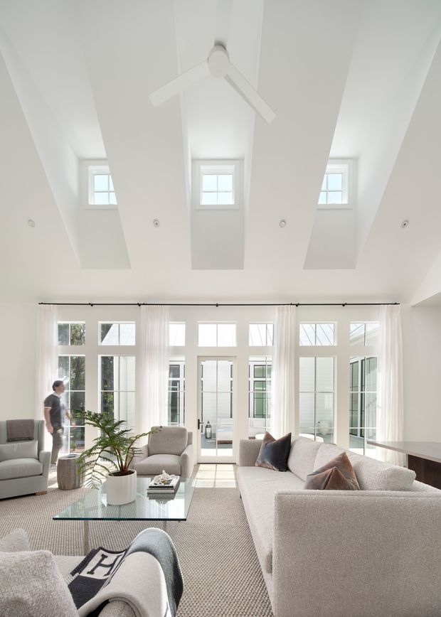 Bright, airy living room with a large light-coloured sectional sofa, matching armchairs, glass coffee table, and neutral rug. Floor-to-ceiling-style glazed doors and windows line the rear wall, while a dramatic vaulted ceiling incorporates multiple projecting skylight-like roof monitors, recessed downlights, and a white ceiling fan. Sheer curtains and restrained furnishings create a refined contemporary interior.