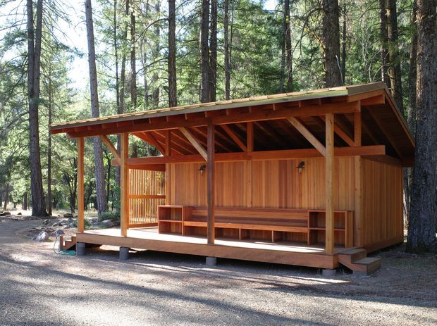 A small open-sided timber pavilion or cabin set among tall pine trees. It has a low-pitched roof with exposed timber rafters and posts, warm vertical timber wall cladding, a raised timber deck, built-in bench seating and shelving, and a partially screened side section. The setting is a quiet forested site with a simple rustic outdoor structure.