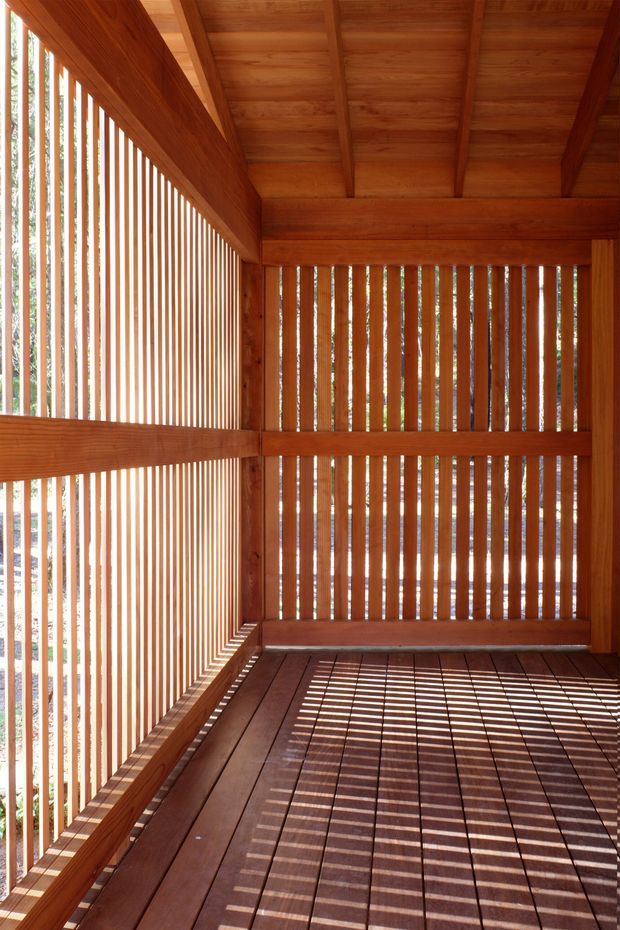 A compact, enclosed timber outdoor space with vertical slatted screens on three sides, a timber-framed overhead roof, and a matching timber plank floor. Strong sunlight creates repeated linear shadows across the floor and walls, with limited views of vegetation and a neighboring structure through the slats.