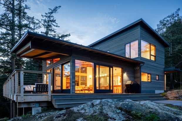 Exterior photograph of a contemporary two-storey home set among dense trees and exposed rocky terrain. The house has dark horizontal cladding, a low-pitched roof with broad overhanging eaves, extensive floor-to-ceiling glazing, and a timber deck with railings. Warm interior lighting is visible through the windows and doors, creating an inviting evening atmosphere.