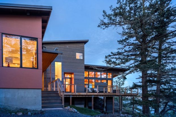 Twilight exterior view of a contemporary multi-level timber-clad house on a sloping, wooded site. The home has broad roof overhangs, large warm-lit windows, a raised timber deck and visible supporting structure beneath, with a mature tree framing the right side.