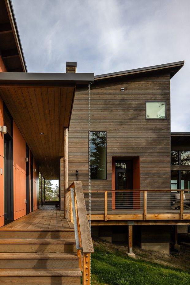 Exterior view of a contemporary multi-level residence with a timber-clad upper volume, broad roof overhangs, covered timber deck and walkway, orange accent walls, and a raised structure supported above a sloping grassy site. The image emphasizes the facade, entry steps, horizontal metal-and-timber balustrades, and a rain chain.