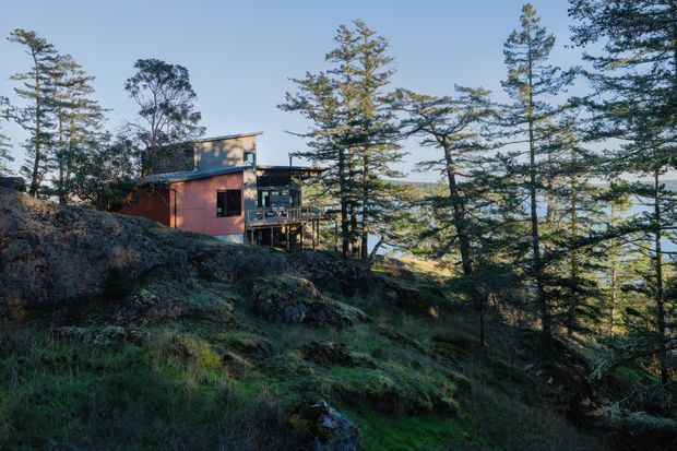 Exterior view of a compact contemporary house set among tall trees on a rocky, moss-covered sloping site. The building has dark timber or metal upper cladding, a muted reddish-brown lower facade, large glazed openings, and projecting deck-like elements. The surrounding forest and rugged terrain dominate the scene.