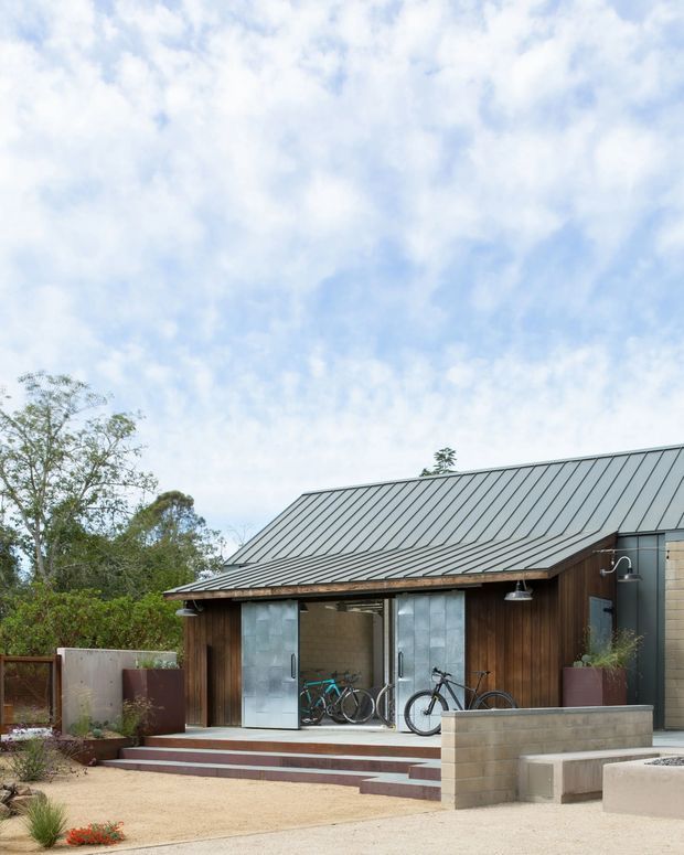 A compact contemporary single-storey building set in landscaped grounds, with a dark standing-seam metal gable roof, vertical timber cladding, and large sliding glazed doors. Bicycles are visible inside the open glazed entry, while concrete block planter walls, steps, and outdoor fixtures define the foreground. The image is primarily an exterior architectural view.