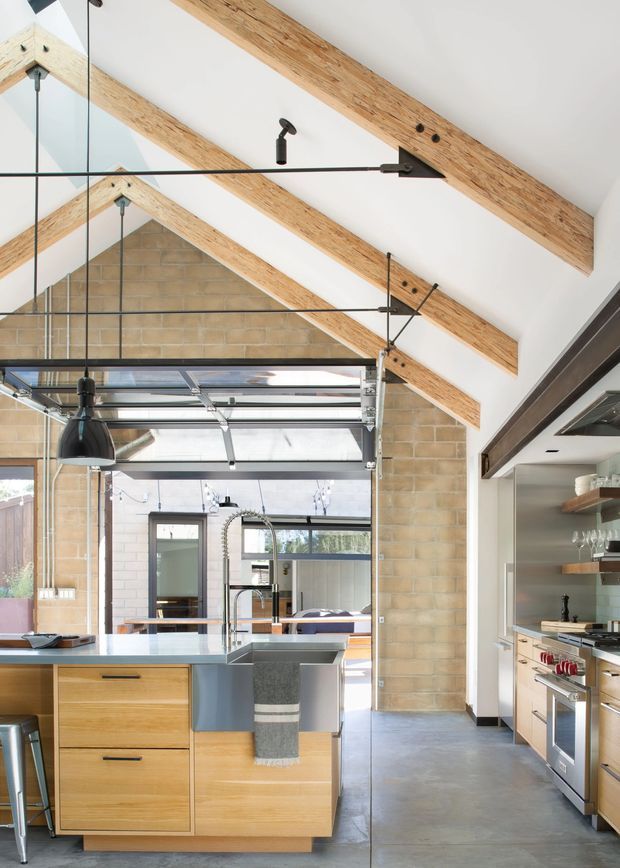 A bright, open-plan kitchen with a central timber-fronted island, pale benchtop, integrated sink and seating. The space features exposed timber roof beams, white vaulted ceilings, tan brick walls, polished concrete flooring, black pendant and track lighting, and large glazed garage-style doors opening to an adjacent area. Fixed kitchen joinery and stainless-steel appliances line the right wall.